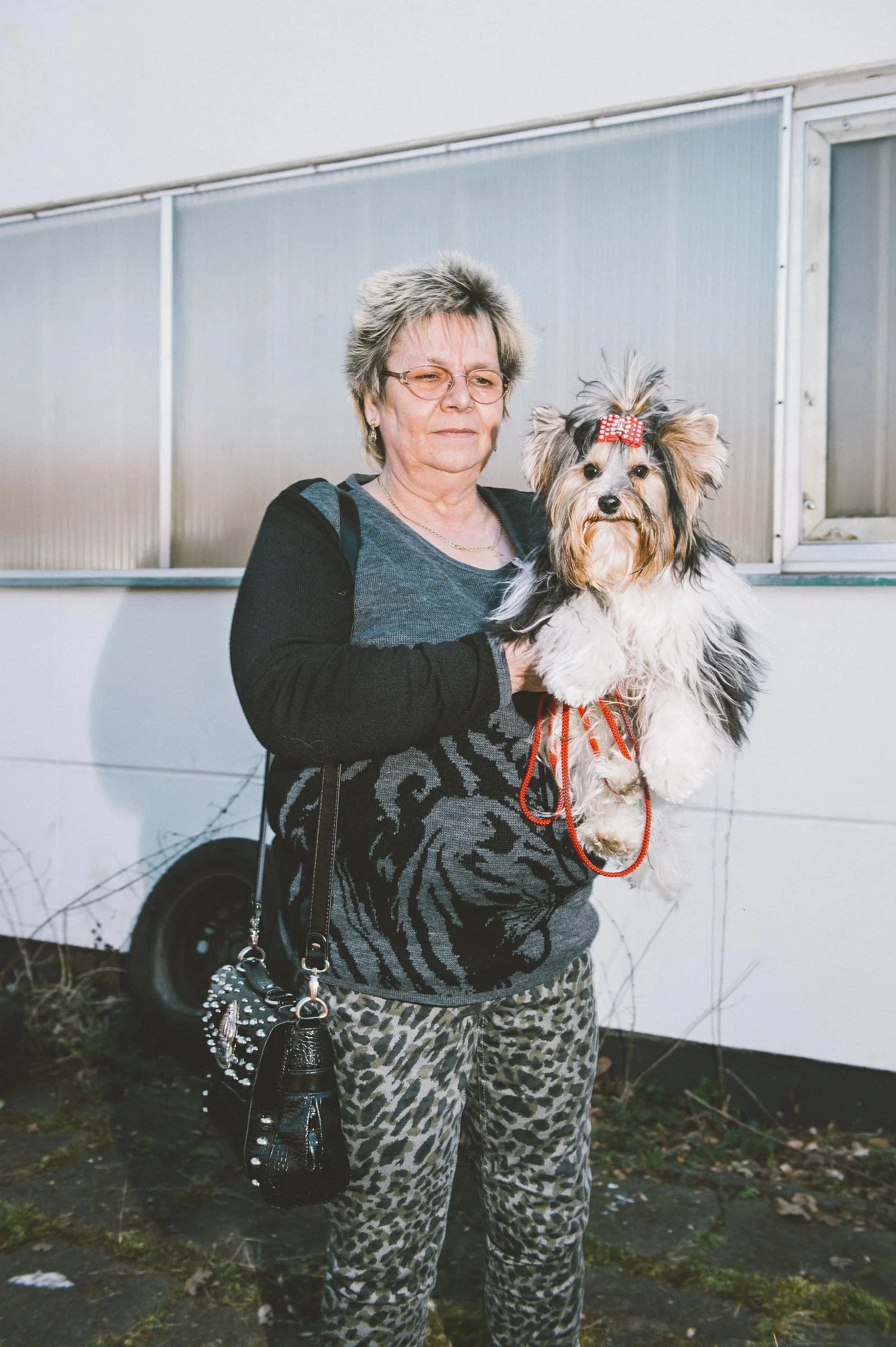 Woman in leopard-print pants proudly holding a small dog with a red bow.